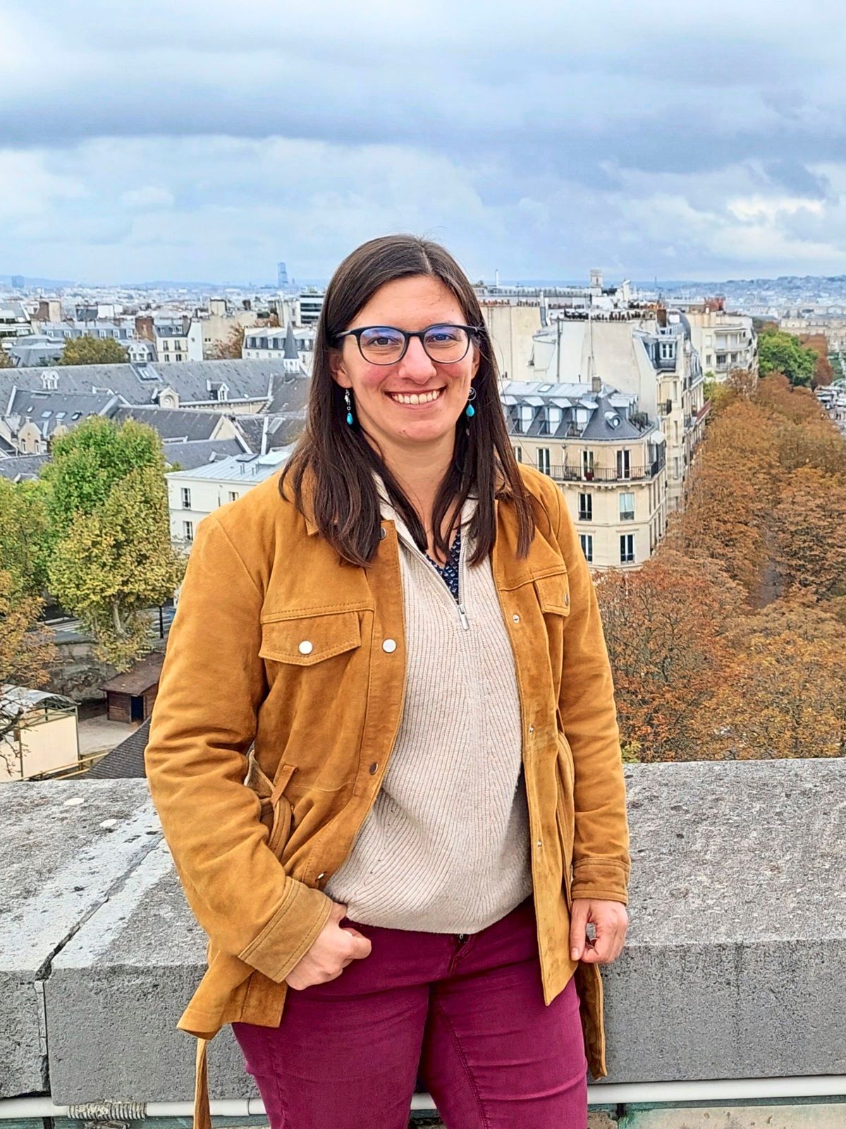 Eine Frau mit langen dunklen Haaren und Brille steht lächelnd auf einer Terrasse mit Blick auf eine Stadtlandschaft. Sie trägt eine hellbraune Jacke, einen cremefarbenen Pullover und bordeauxrote Hosen. Im Hintergrund sind Gebäude, Bäume, und ein bewölkter Himmel zu sehen.