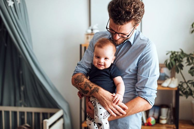 Ein Vater steht in einem Kinderzimmer mit Gitterbett, ein Kleinkind auf dem Arm