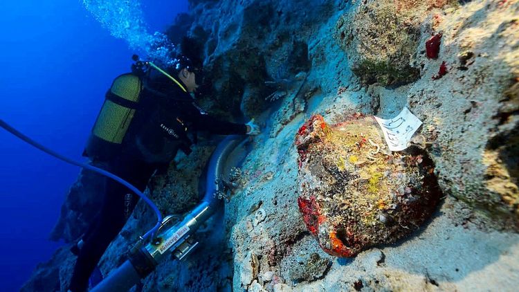 Ein Taucher untersucht unter Wasser die Überreste eines antiken Schiffswracks. Im Vordergrund ist eine Amphore aus der frühen Bronzezeit zu sehen, die teilweise mit Meeresbewuchs bedeckt ist. Ein Schild mit einer Nummer ist an der Amphore befestigt. Im Hintergrund sind Felsen und Ausrüstung zu erkennen.