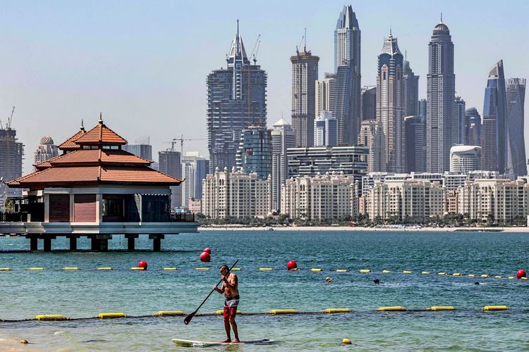 Ein Mann übt Stand-up-Paddleboarding (SUP) im Wasser vor dem Anantara The Palm Dubai Resort. Im Hintergrund sind die Skyline von Dubai mit hohen Wolkenkratzern und ein Gebäude im traditionellen Stil zu sehen.