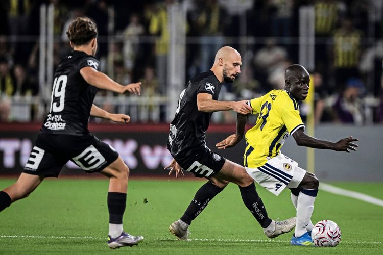 Ein Fußballspiel zwischen Fenerbahçe und Sturm Graz: Ein Spieler von Fenerbahçe im gelb-blauen Trikot dribbelt den Ball, während zwei Spieler von Sturm Graz in schwarzen Trikots versuchen, ihn zu stoppen. Im Hintergrund sind Zuschauer und das Spielfeld zu sehen.