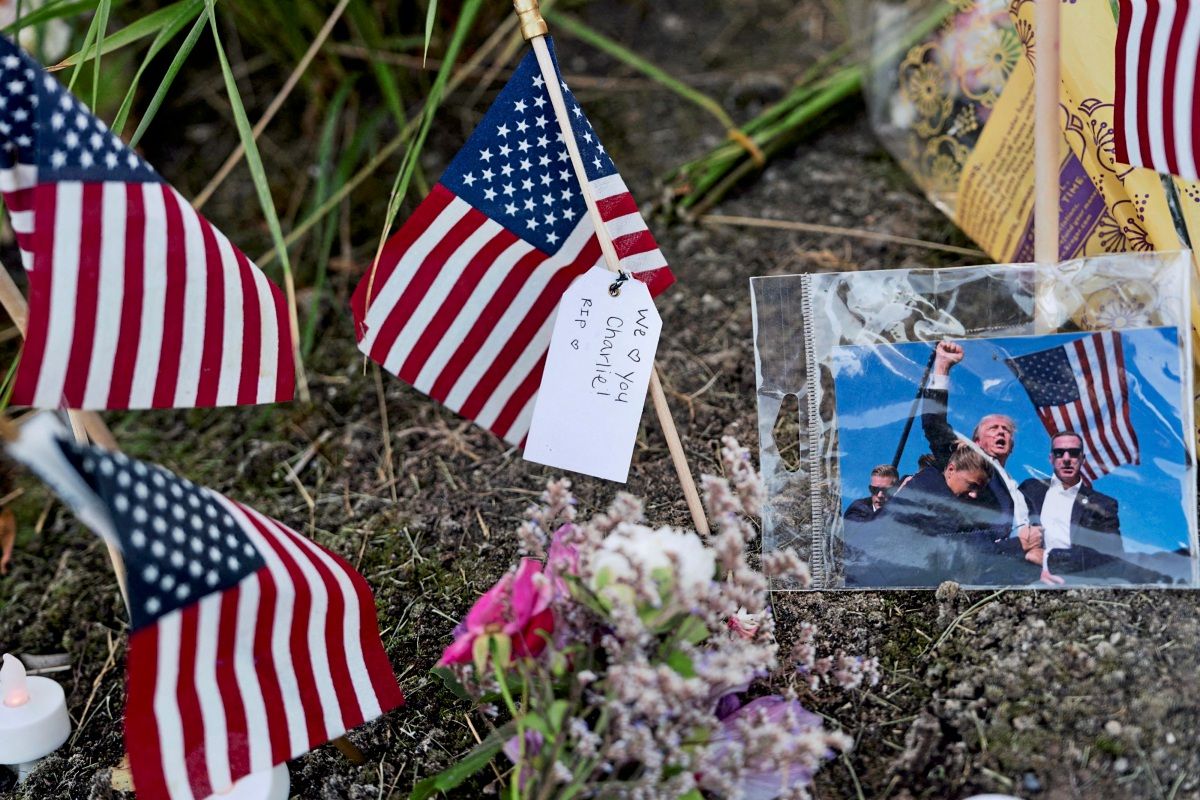 Three American flags and the image of the failed assassination attempt on Donald Trump in Butler during the 2024 campaign lie on the ground next to some flowers.