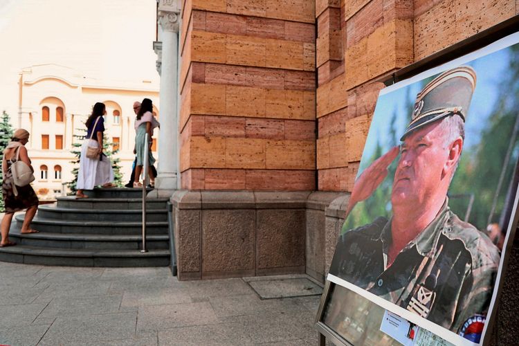 Eine Fotografie zeigt ein großes Poster mit einem Porträt von Ratko Mladic in Militäruniform, ausgestellt vor der orthodoxen Kathedrale in Banja Luka, Bosnien. Im Hintergrund betreten mehrere Personen die Treppe des Gebäudes.