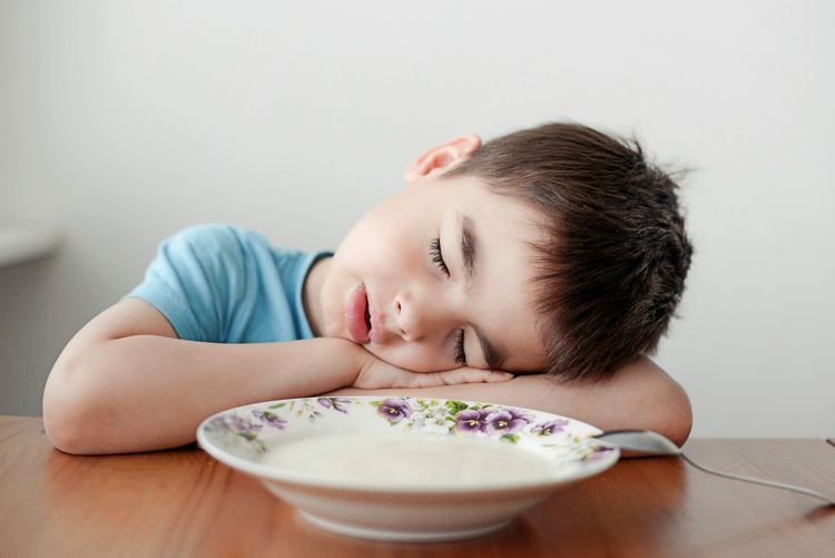 Ein Kind in einem blauen T-Shirt hat beide Arme auf einen Tisch und den Kopf seitlich daraufgelegt, vor sich eine halbvolle Schüssel mit Blumenmuster, gefüllt mit einer Art Suppe. Es hat die Augen müde und erschöpft geschlossen und den Mund leicht geöffnet.