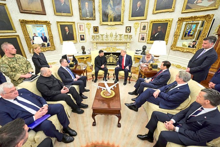 Eine Gruppe von Personen, darunter der ukrainische Präsident Wolodymyr Selenskyj (links in der Mitte) und der US-Präsident Donald Trump (rechts in der Mitte), sitzt in einem elegant eingerichteten Raum mit goldenen Akzenten und vielen Porträts an den Wänden. Gesprächsatmosphäre in einer offiziellen Besprechung. Im Vordergrund ein Tisch mit einem Modellflugzeug.