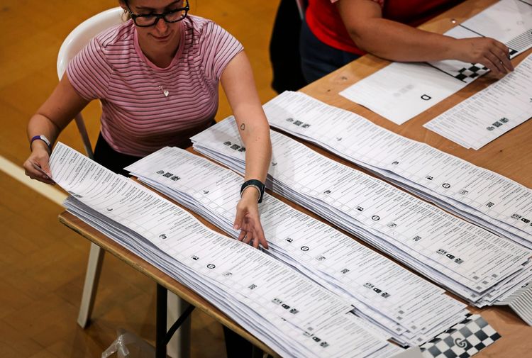 Eine Frau zählt lange Stimmzettel, die in mehreren Stapeln auf einem Tisch liegen. Sie trägt ein gestreiftes rosa Oberteil und eine Brille. Im Hintergrund ist eine weitere Person zu sehen.