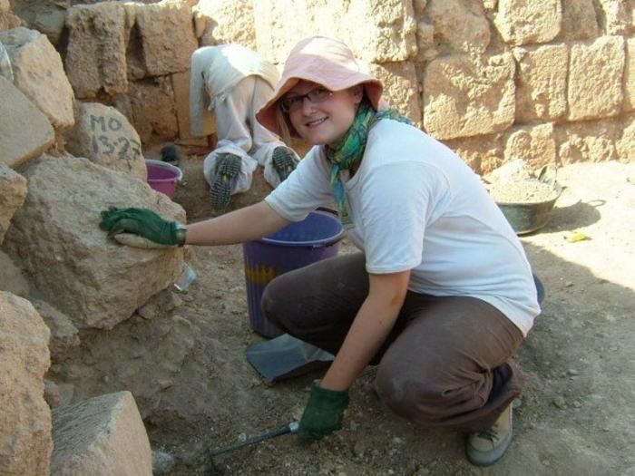 Eine Archäologin mit Sonnenhut mit Schutzhandschuhen vor einer Steinmauer