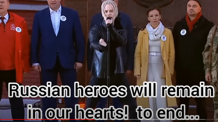 Zu sehen ist der Sänger Shaman am Mikrofon vor hinter ihm stehenden russischen Honoratioren.