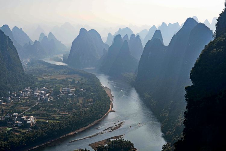 Eine Luftaufnahme zeigt den Li-Fluss, der sich durch markante, nebelverhangene Karsthügel schlängelt, nahe Yangshuo in der Provinz Guangxi, China. Am Ufer sind kleine Dörfer und Felder zu sehen.