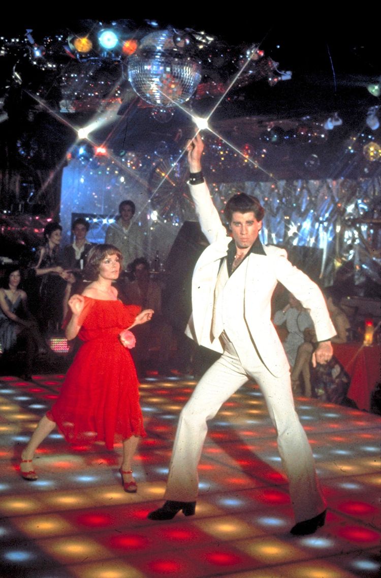 Ein Mann in einem weißen Anzug tanzt mit einer Frau in einem roten Kleid auf einer beleuchteten Disco-Tanzfläche. Im Hintergrund sind eine Discokugel und Zuschauer zu sehen.