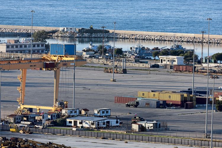 Eine Übersicht über eine russische Marineeinrichtung in Tartus, an Syriens Küste. Im Vordergrund sind ein Hafen mit Containern, gelben Kränen und kleinen Gebäuden zu sehen. Im Hintergrund liegt das Meer mit Booten und einer Hafenmauer.