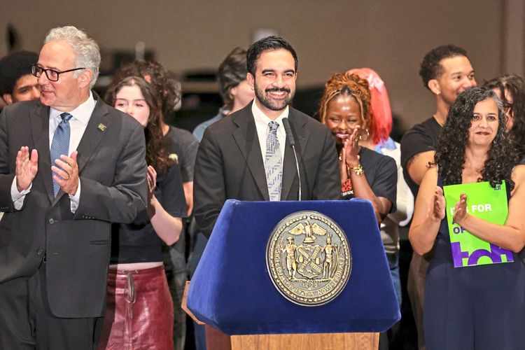 Eine Gruppe von Menschen steht bei einer Pressekonferenz auf einer Bühne. Im Mittelpunkt steht ein Mann im Anzug, der an einem Rednerpult mit dem Siegel von New York spricht und lächelt. Personen auf beiden Seiten applaudieren, einige halten Plakate. Im Hintergrund sind weitere Personen zu sehen.