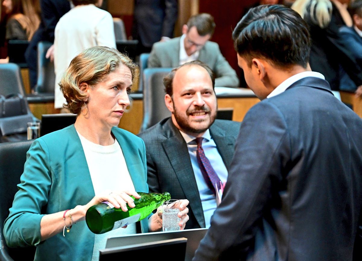 Drei Personen in einem Gespräch während einer Sitzung des österreichischen Nationalrates im Parlament in Wien. Eine Frau gießt Wasser in ein Glas, während zwei Männer mit ihr sprechen. Im Hintergrund sind weitere Personen und Sitzplätze zu sehen.
