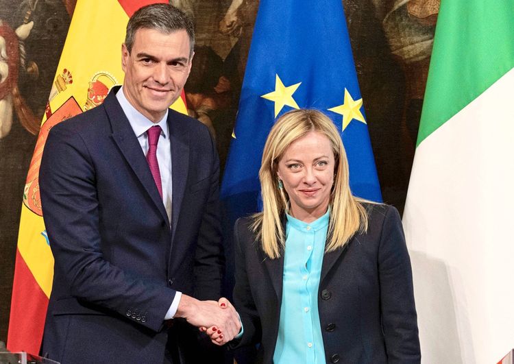 Zwei Personen stehen vor den Flaggen von Spanien, der Europäischen Union und Italien. Sie geben sich die Hand und blicken in die Kamera.