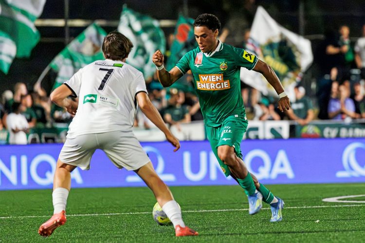Ein Fußballspielbild: Ein Spieler im grünen Trikot des SK Rapid dribbelt mit dem Ball, während ein Spieler des SV Wienerberg im weißen Trikot versucht, ihn zu verteidigen. Im Hintergrund sind Zuschauer und Vereinsfahnen zu sehen.