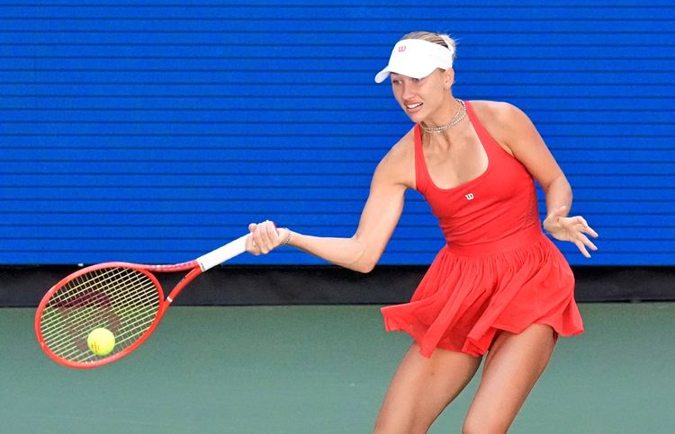 Eine Tennisspielerin in einem roten Outfit und mit weißem Visor schlägt während eines Spiels den Ball mit ihrem Schläger. Im Hintergrund ist eine blaue Wand zu sehen.