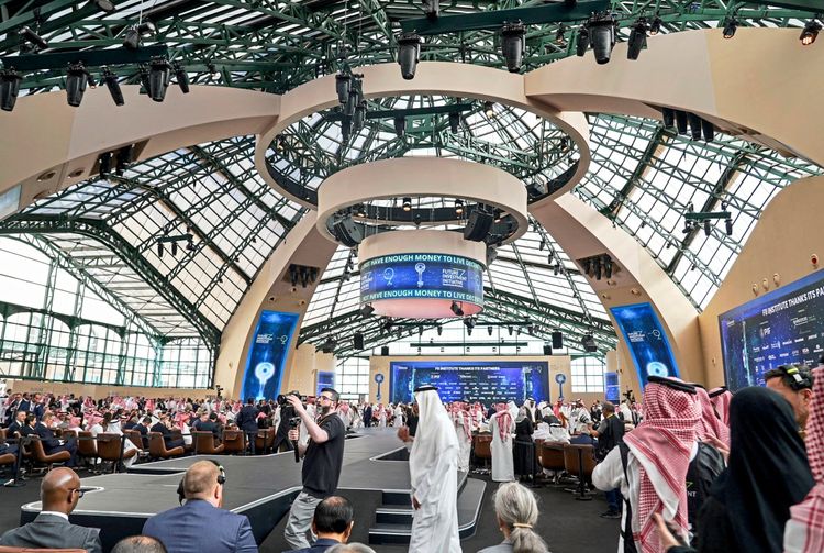 Eine große Veranstaltungshalle mit einer modernen Glas- und Stahlarchitektur, gefüllt mit Teilnehmern in formeller Kleidung, darunter traditionelle saudi-arabische Gewänder. Im Vordergrund ist eine Bühne zu sehen, während im Hintergrund Bildschirme mit Konferenzdetails angezeigt werden. Die Szene zeigt die 9. Ausgabe der Future Investment Initiative (FII) 2025 in Riad, Saudi-Arabien.