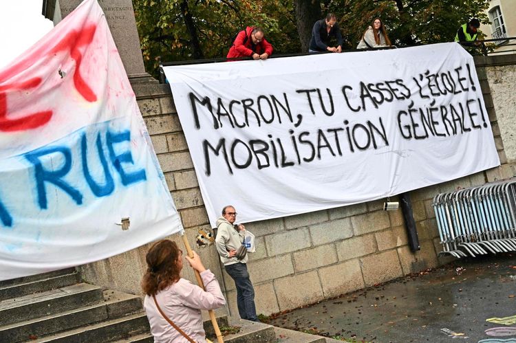 Eine Gruppe von Menschen befestigt ein großes weißes Banner an einer Wand mit der Aufschrift „MACRON, TU CASSES L'ÉCOLE! MOBILISATION GÉNÉRALE!“ während einer Demonstration in Nantes, Frankreich, am 13. Oktober 2026. Im Vordergrund schwenkt eine Person eine andere Fahne mit Schriftzügen in Rot und Blau. Die Proteste richten sich gegen schlechte Lernbedingungen an Schulen.