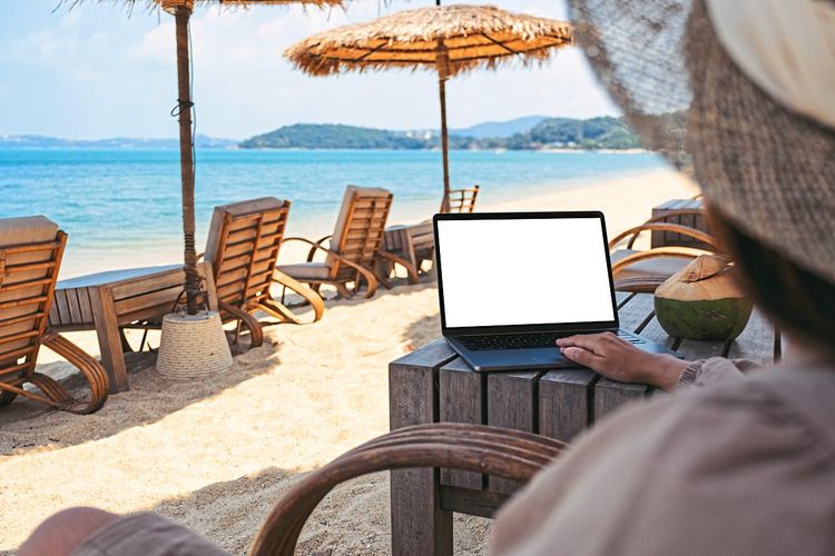 Frau mit Sonnenhut sitzt mit Laptop auf einer Strandliege mit Blick aufs Meer