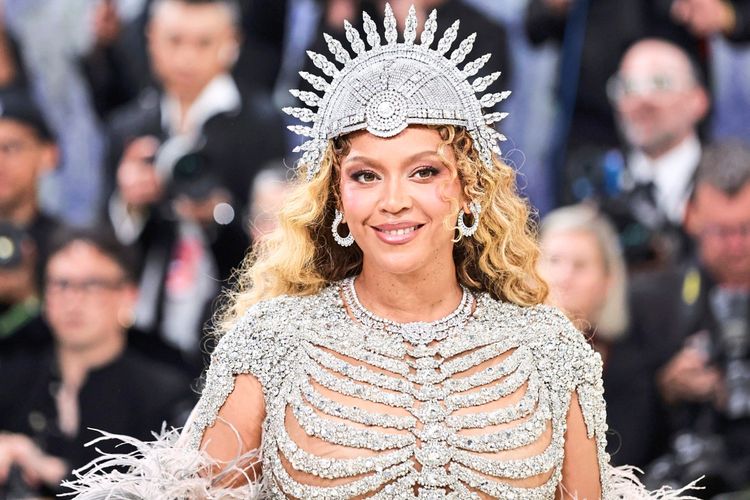 Bildbeschreibung: Eine Frau mit lockigen, blonden Haaren trägt eine silberne, glitzernde Krone und ein funkelndes Outfit mit Skelett-Design. Sie lächelt auf dem roten Teppich und befindet sich bei einer glamourösen Veranstaltung. Im Hintergrund sind unscharfe Personen zu sehen.