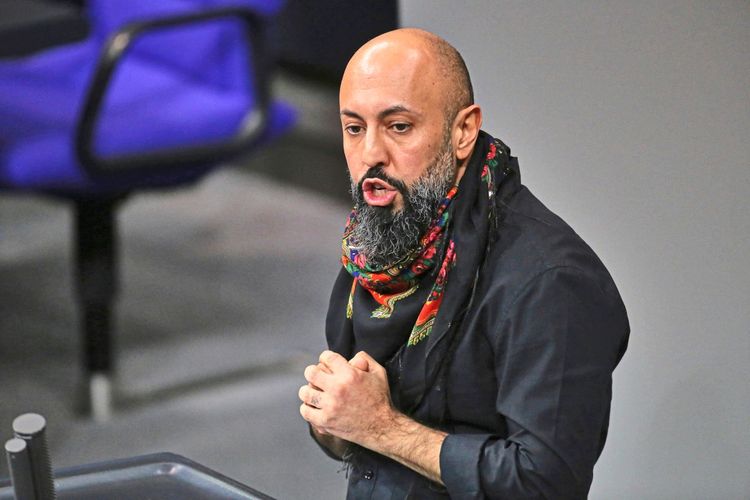 Ein Mann mit Glatze und dichtem Bart, bekleidet mit einem schwarzen Hemd und einem bunten Schal, spricht leidenschaftlich in einem Raum, der wie ein Plenarsaal aussieht. Im Hintergrund ist ein blauer Stuhl sichtbar.