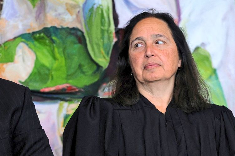 Eine Frau in schwarzer Robe sitzt vor einem farbenfrohen, abstrakten Hintergrund und blickt zur Seite.