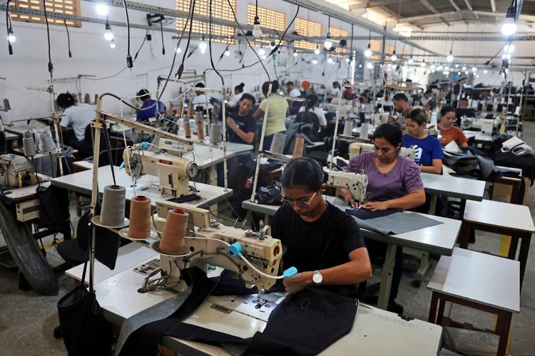 Arbeiterinnen und Arbeiter sitzen an Nähmaschinen in einer hell beleuchteten Textilfabrik in Lajes, Brasilien. Sie nähen Kleidung und arbeiten konzentriert. Im Hintergrund sind weitere Personen und Maschinen zu sehen.