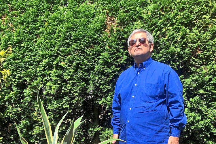 Ein Mann steht in einem sonnigen Garten auf einer gepflasterten Fläche vor einer hohen Hecke. Er trägt ein blaues Hemd, eine beige Hose und dunkle Sportschuhe. Neben ihm steht ein Holzstuhl mit einer großen Pflanze in einem Topf.