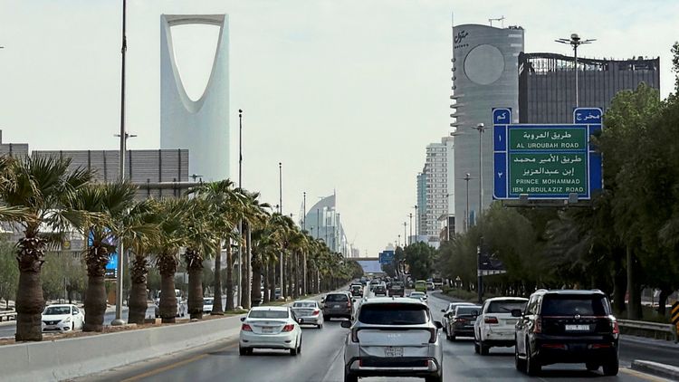 Straßenansicht in Riad, Saudi-Arabien, mit viel Verkehr, modernen Gebäuden, darunter dem markanten Kingdom Tower, Palmen entlang der Straße und Straßenschildern in Arabisch und Englisch.