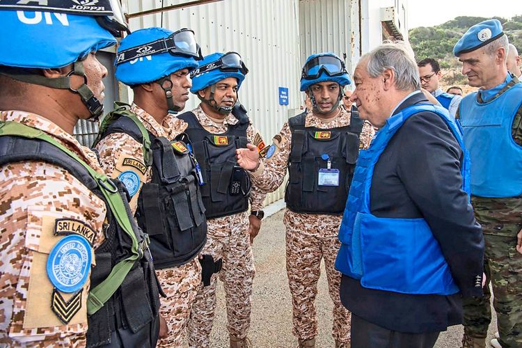 Eine Gruppe von UN-Friedenssoldaten mit blauen Helmen unterhält sich mit einem Mann in einem blauen Schutzanzug, begleitet von anderen Personen. Im Hintergrund ist ein UNIFIL-Hauptquartier in Naqura, Südlibanon, nahe der israelischen Grenze, zu sehen.