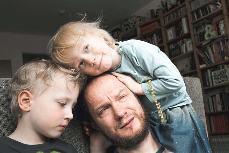 Vater mit zwei kleinen Kindern sitzt im Wohnzimmer, eines der Kinder klettert auf seinen Kopf, er zwickt die Augen zusammen