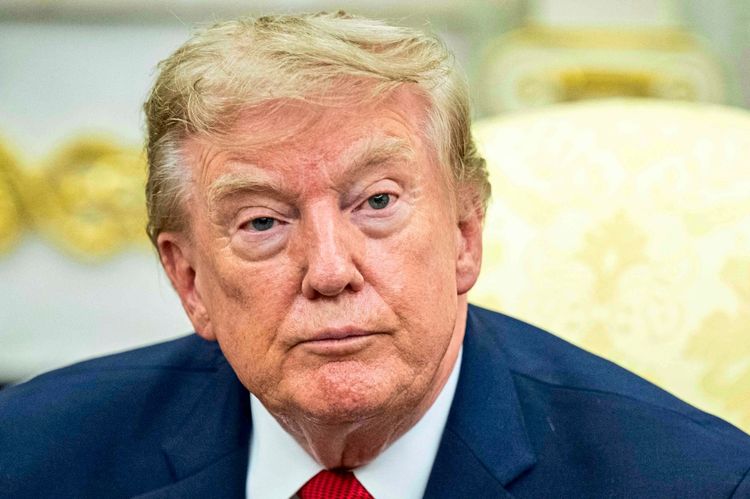 Ein Mann mit blondem Haar in einem dunklen Anzug sitzt vor einem goldenen Hintergrund und blickt nachdenklich.