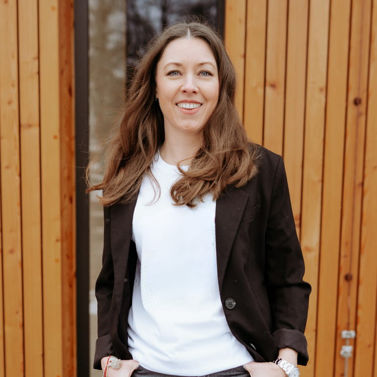 Eine Frau mit langen, braunen Haaren steht lächelnd vor einer Holzverkleidung. Sie trägt ein weißes T-Shirt, einen schwarzen Blazer und eine Uhr am Handgelenk.