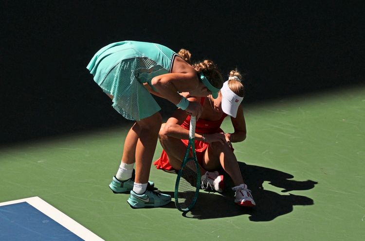 Zwei Tennisspielerinnen auf einem Tennisplatz: Eine Spielerin in einem türkisfarbenen Outfit beugt sich besorgt zu einer anderen Spielerin in einem roten Outfit, die auf dem Boden sitzt, nachdem sie sich verletzt hat.