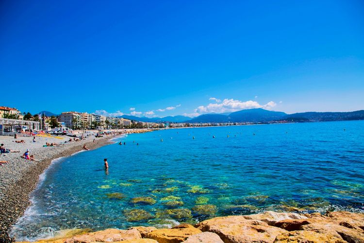 Strand und azurblaues Meer an der Côte d'Azur in Nizza, Frankreich, mit Bergen und Gebäuden im Hintergrund. Menschen entspannen und schwimmen am Kiesstrand unter klarem Himmel.