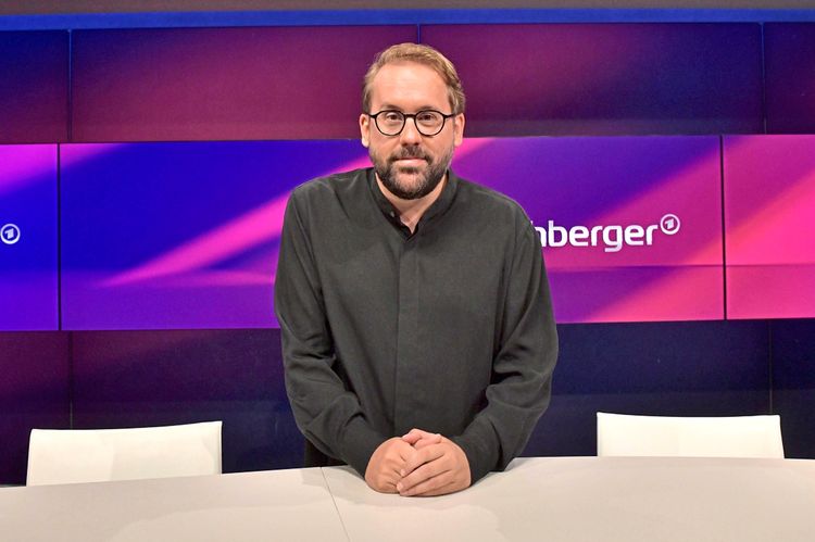 Ein Mann mit Brille und Bart steht an einem Tisch in einem Studio. Im Hintergrund ist ein Bildschirm mit dem Schriftzug 