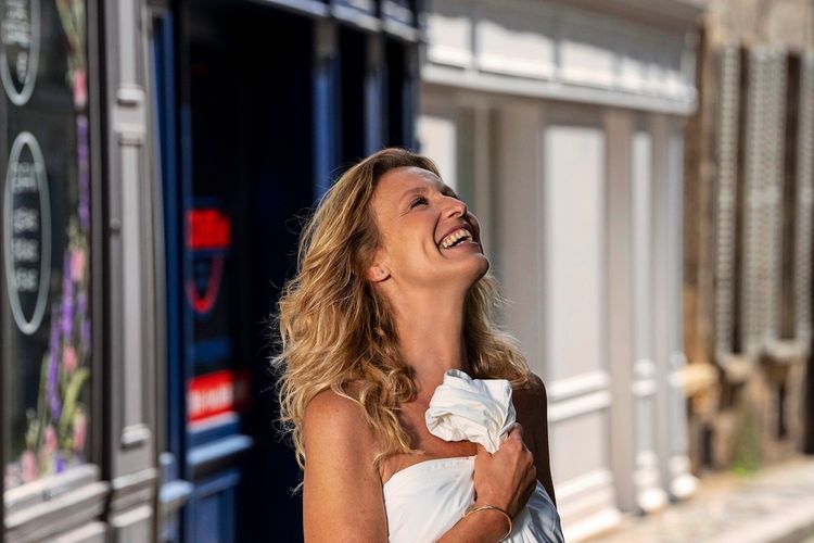 Eine Frau mit blonden Haaren lacht fröhlich und hält ein weißes Tuch in der Hand. Sie befindet sich in einer städtischen Umgebung mit Gebäuden im Hintergrund.