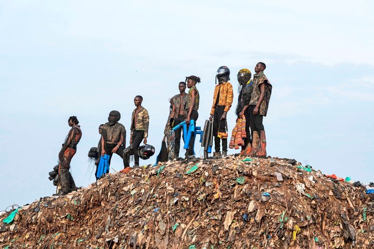 Eine Gruppe von Menschen steht auf einem großen Müllberg. Sie tragen künstlerische, kreative Kleidung und Accessoires, darunter Helme und farbenfrohe Materialien, und blicken in verschiedene Richtungen. Der Himmel im Hintergrund ist hell und wolkig.