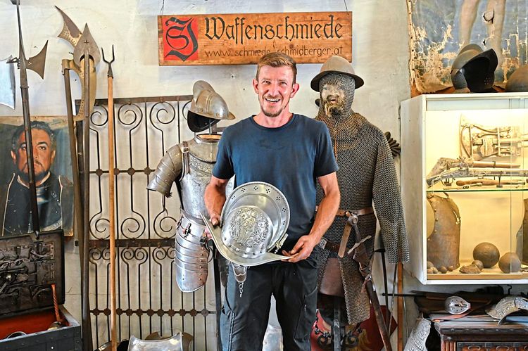 Der Schmied Johann Schmidberger mit einem Helm in der Hand, hinter ihm ein Harnisch und eine alte Rüstung.
