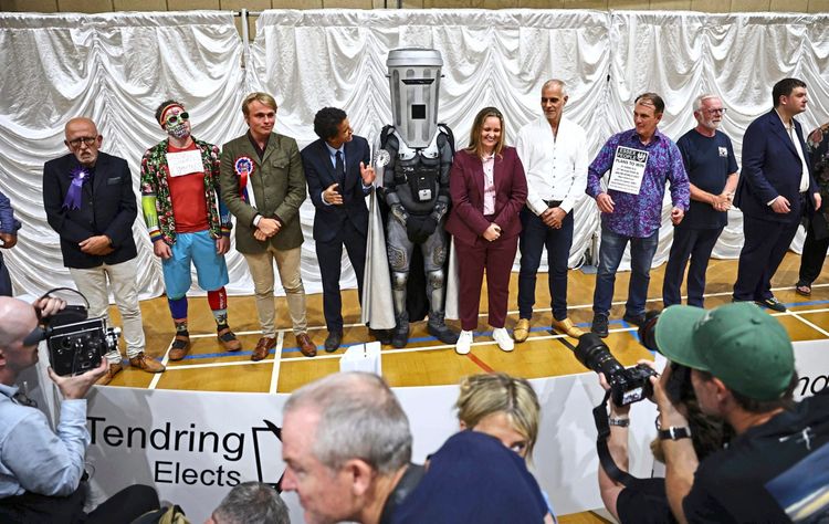 Eine Gruppe von Kandidaten steht nebeneinander in einer Halle mit weißen Vorhängen, während Kameras sie fotografieren. Unter ihnen ist eine Person in einem auffälligen Metallic-Kostüm (