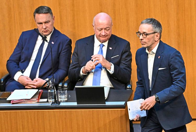 Drei Männer in Anzügen sitzen bzw. stehen in einem Sitzungssaal. Zwei Männer sitzen nebeneinander an einem Tisch mit Mikrofonen und Wasserflaschen, während der Mann rechts, mit Papieren in der Hand, vorbei geht. Hintergrund aus Holzverkleidung.