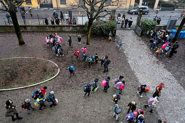Luftaufnahme einer Grundschule: Kinder mit Rucksäcken stehen in kleinen Gruppen auf einem Schulhof, umgeben von kahlen Bäumen und Gehwegen. Im Hintergrund ist ein Eingangstor, wo Erwachsene warten. Die Szene zeigt den Schulbeginn unter COVID-19-Regelungen in Italien.
