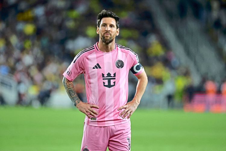 Ein Fußballspieler in einem rosa Trikot von Inter Miami CF steht auf einem Spielfeld. Er trägt eine Kapitänsbinde am linken Arm und hat die Hände in die Hüften gestützt. Im Hintergrund ist das unscharfe Stadion mit Zuschauern zu sehen.