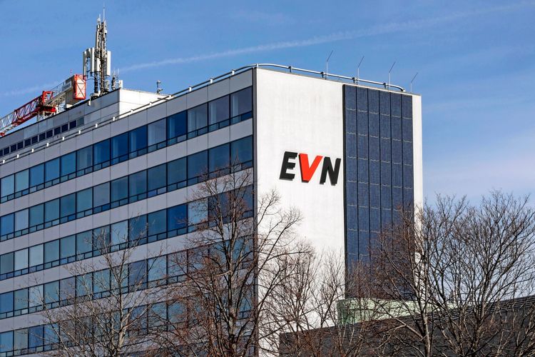 Das Bild zeigt das Hauptgebäude der EVN in Maria Enzersdorf, Niederösterreich, Österreich. Es handelt sich um ein weißes, modernes Gebäude mit großen Fensterfronten und einem vertikalen Abschnitt aus Solarpanelen. Oben auf dem Gebäude befinden sich Antennen und eine Baustellenkrananlage. Im Vordergrund sind kahle Baumkronen sichtbar.
