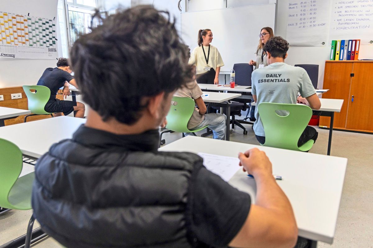Eine Unterrichtssituation in einem Klassenraum. Jugendliche sitzen an Tischen, während zwei Frauen vorne stehen und unterrichten. Im Hintergrund sind eine Tafel, ein Wandkalender und Regale mit Ordnern zu sehen.