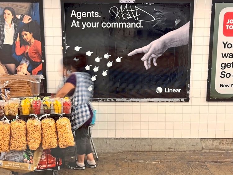 Eine Person steht an einem Verkaufswagen in einer U-Bahn-Station. Der Wagen ist mit Lebensmitteln wie Chips, Süßigkeiten und Getränken beladen. Im Hintergrund befinden sich Werbeanzeigen an einer gekachelten Wand.