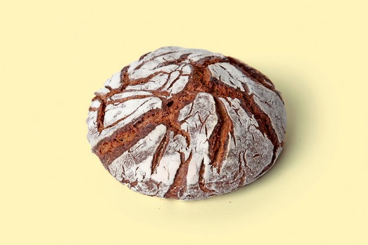 Ein rundes Roggenbrot mit rissiger, bemehlter Kruste vor einem hellgelben Hintergrund.