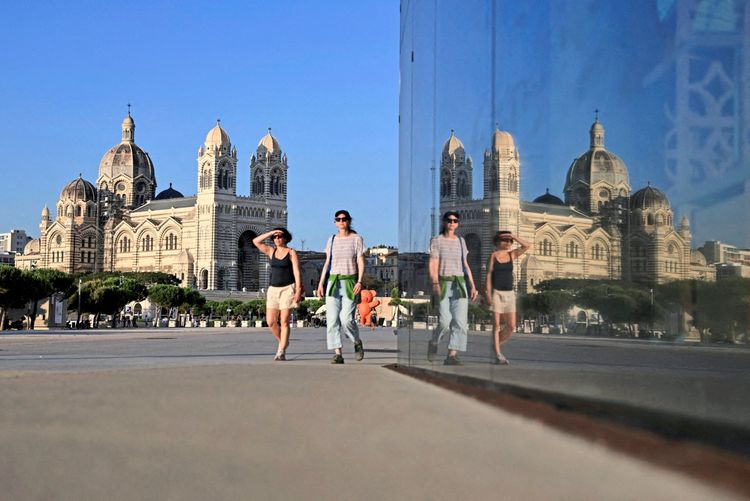 Zwei Frauen gehen an einer Glasfläche vorbei, die die Kathedrale La Major in Marseille, Frankreich, reflektiert. Der Himmel ist klar und blau.