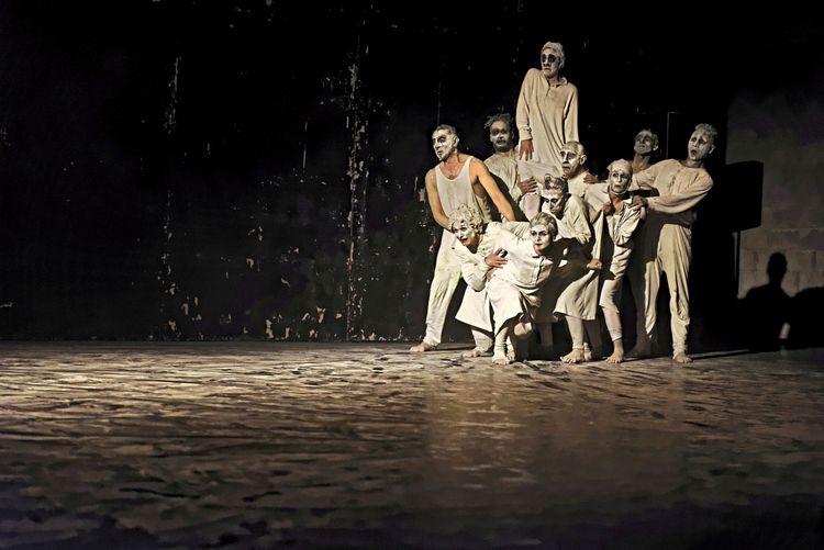 Eine Gruppe von Darstellern in weißen Kostümen und mit weißer Gesichtsbemalung steht auf einer dunklen Bühne. Sie posieren eng beieinander und wirken in eine Performance vertieft. Die Beleuchtung betont ihre Gestalten und hebt sie vom dunklen Hintergrund ab.