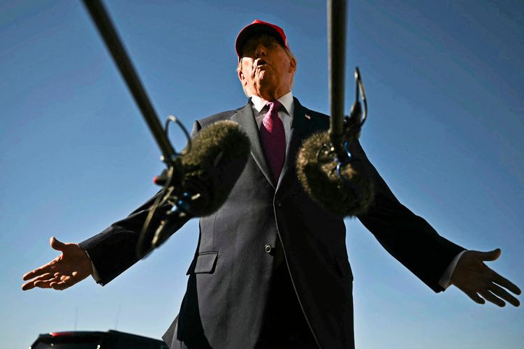 Donald Trump in einem Anzug und roter Kappe spricht mit weit geöffneten Armen in Mikrofone. Der Himmel im Hintergrund ist klar und blau.